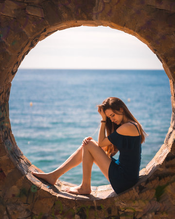 A young attractive woman from Spain in a blue dress sitting in the stone round window by the seaの写真素材