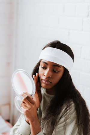 An attractive young African female with long hair applying face cream while looking in a hand mirror in the bedroomの写真素材