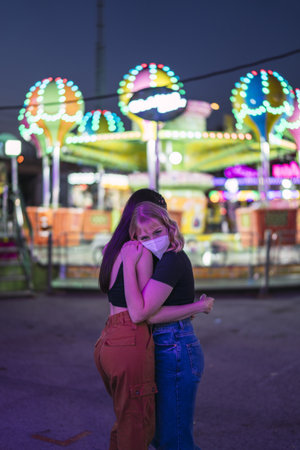 Two cheerful Caucasian female friends with masks hugging at an amusement park in the eveningの写真素材