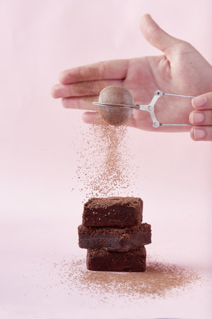 A closeup shot of hands powdering cacao powder on a piece of freshly baked chocolate brownie cakeの写真素材