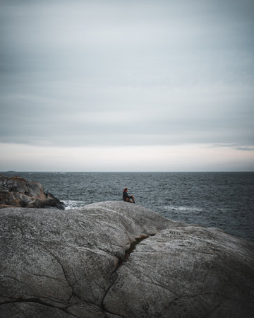 A vertical shot of a man sitting on the cliff looking at the wavy sea against cloudy sky backgroundの写真素材