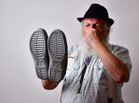 A senior Caucasian bearded male covering his nose while holding stinky shoes