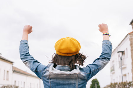 A back view of a female in a yellow cap and denim jacket with hands up; an LGBT bracelet on the right handの写真素材