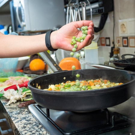 A hand pouring peas over vegetables frying in a small amount of fat in the panの写真素材