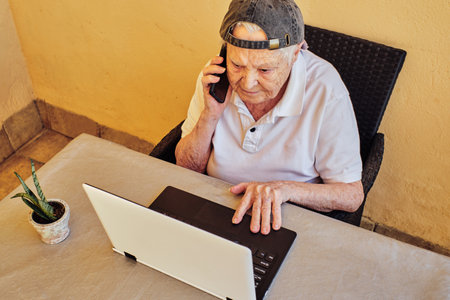 A thoughtful elderly female talking on the phone while working with a laptopの写真素材
