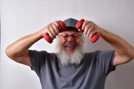 A happy American man with glasses and cap holding dumbbells and training on a white wall background