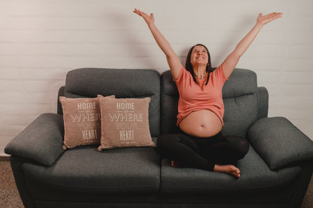 A Caucaisan pregnant woman sitting on sofa with raised arms - happiness during pregnancy conceptの写真素材