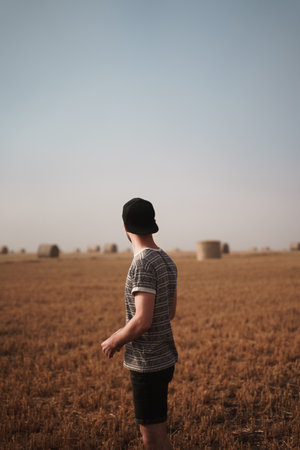 A back view of a male in a gray shirt and a hat standing on the grassy field under the blue skyの写真素材