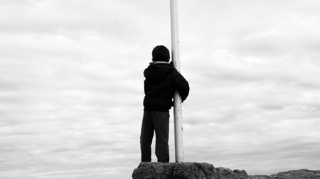 A grayscale shot of a child hugging a pole under a cloudy skyの写真素材