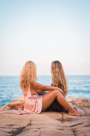 A couple of attractive sexy caucasian girls in summer dresses sitting on orange rocks and watching the seaの写真素材