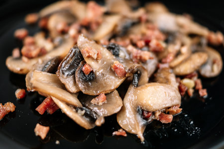 A closeup shot of cooked tasty mushrooms with ham pieces on a black plateの写真素材