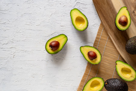 A top view of fresh halved avocados and a wooden cutting board on a white surface, copy space