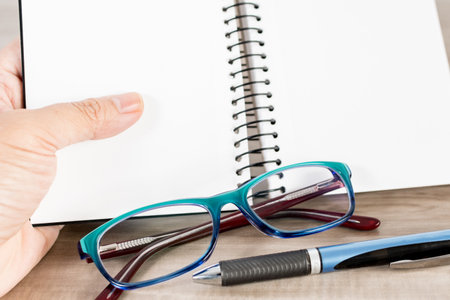 A female's hand holding a notebook with a pencil and glasses on a wooden table