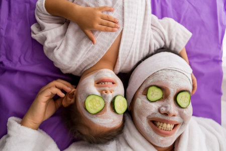 A mother and her young pretty daughter having fun in the bedroom and making clay facial masks with cucumber covering their eyesの写真素材