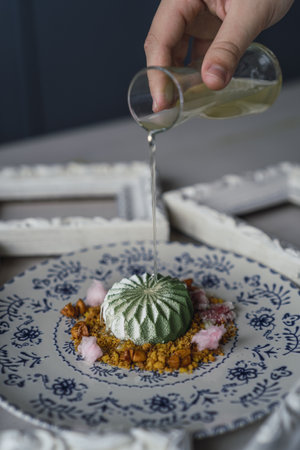 A vertical shot of a chef pouring sweet sauce on a small green cake with biscuit crumbles on a plateの写真素材