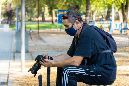 Stock photo of man wearing face mask looking at some photos in his camera while sitting in the park.
