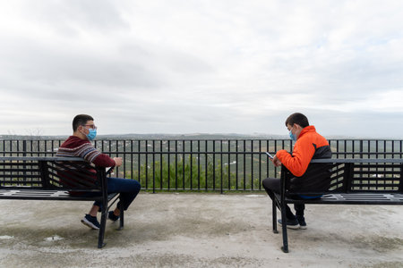Stock photo of teenagers wearing face mask due to covid19 sitting in a bench and using phone.