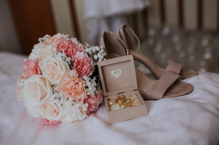 A selective focus shot of golden wedding rings, bridal bouquet, and heels on white sheets