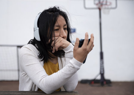 A beautiful Hispanic woman with headphones watching a movie on a phone outdoorsの写真素材