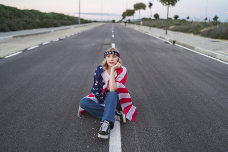 A young Caucasian female sitting on the asphalt road with the flag of the USA on her shouldersの写真素材