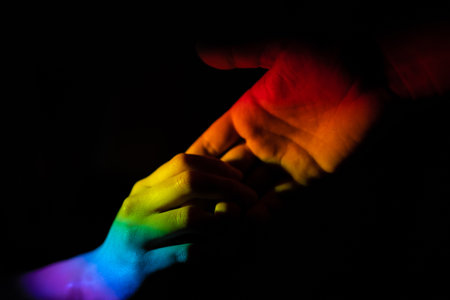 A closeup of a hand helping another illuminated with the LGBT pride flag on black background - love...