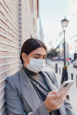 A Caucasian female in a white protective mask using her smartphone while leaning on the wallの写真素材