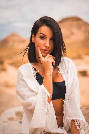 A sensual look of a young brunette Caucasian girl in a white dress sitting in a beautiful desertの写真素材