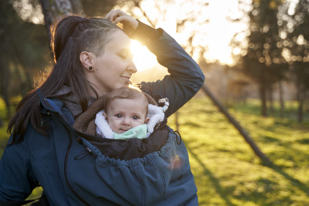 A beautiful view of the mother carrying her child in front of her in the coat on a sunny day outside