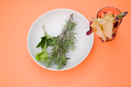 A closeup of a plate with rosemary branch and greens next to glass with iris flower on peach surfaceの写真素材