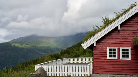 A stunning view of clouds and rainbow on a valley from a traditional red, Norwegian cottageの写真素材