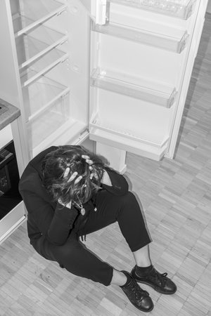 Depressed Woman Sitting on the Floor in Front of an Empty Refrigerator at Home in Switzerland.の写真素材