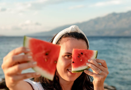 A portrait of a young Croatian female holding two watermelon slices while having fun at the beachの写真素材