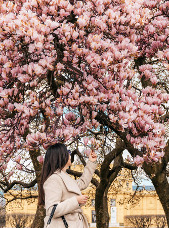 Young woman standing in park in city, touching pink blossom of magnolia tree.
