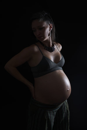 A beautiful young pregnant lady posing in a sports bra and a necklace on a black backgroundの写真素材