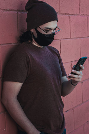 A vertical shot of a handsome caucasian male in glasses and facial mask looking at his phone - new normal conceptの写真素材