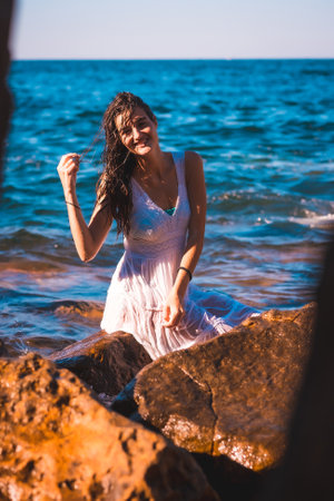 A young Caucasian girl smiling in a white dress and wet hair sitting on a rock by the seaの写真素材