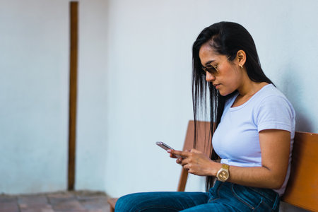 A beautiful young Colombian female in sunglasses sitting on a bench and checking her phone