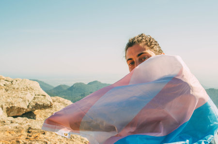 A young Spanish man with the trans flag on his shoulders on a peak of a mountainの写真素材