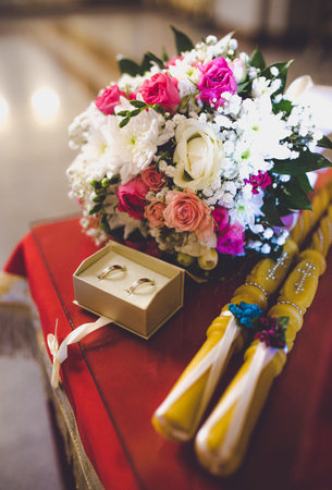 A closeup shot of decorated wedding rings with a bridal bouquet and candles for a Christian wedding