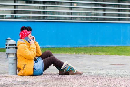A young Caucasian female in casual clothing sitting on a sidewalk and talking on a phoneの写真素材