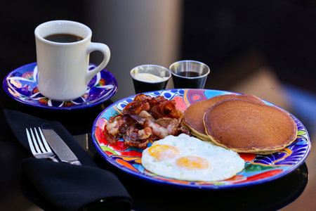 A plate of fried eggs served with bacon and pancakes with a cup of coffee on the tableの写真素材