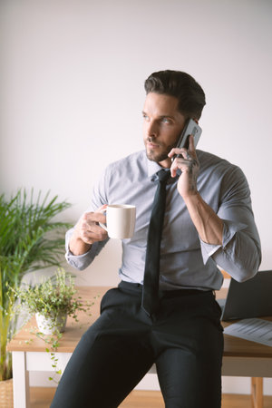A vertical shot of a young businessman drinking coffee and talking on the phone at the officeの写真素材