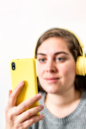 A young woman wearing wireless yellow headphones listens and watches smartphone content sitting at home. Selective focus. Side view.の写真素材