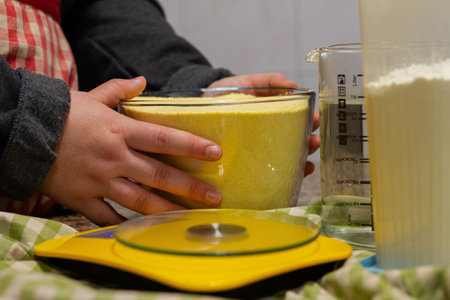 A closeup of a baker holding a bowl of yellow flour to weigh on a scale