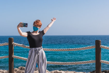 A young caucasian female in a face mask taking a selfie at the beachの写真素材