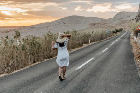 An attractive Croatian woman in a white dress walking on a narrow road through a valley during sunsetの写真素材