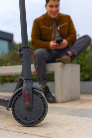 An electric scooter in the background of a young Hispanic man resting on a bench and using a...