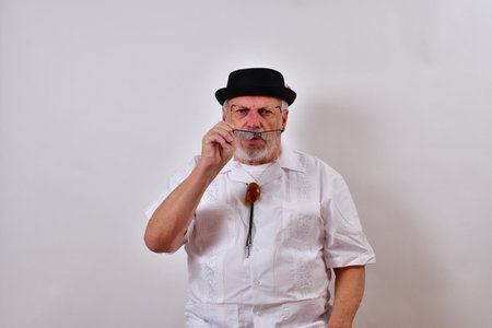 A view of an elderly man wearing a hat and a white shirt trying to put on his glasses on whiteの写真素材