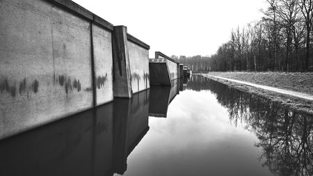 A black and white shot of a canal with tree reflections and a concrete wallの写真素材