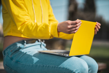 A closeup shot of a girl's hands adjusting her yellow laptop screen balanced on her knees in the parkの写真素材